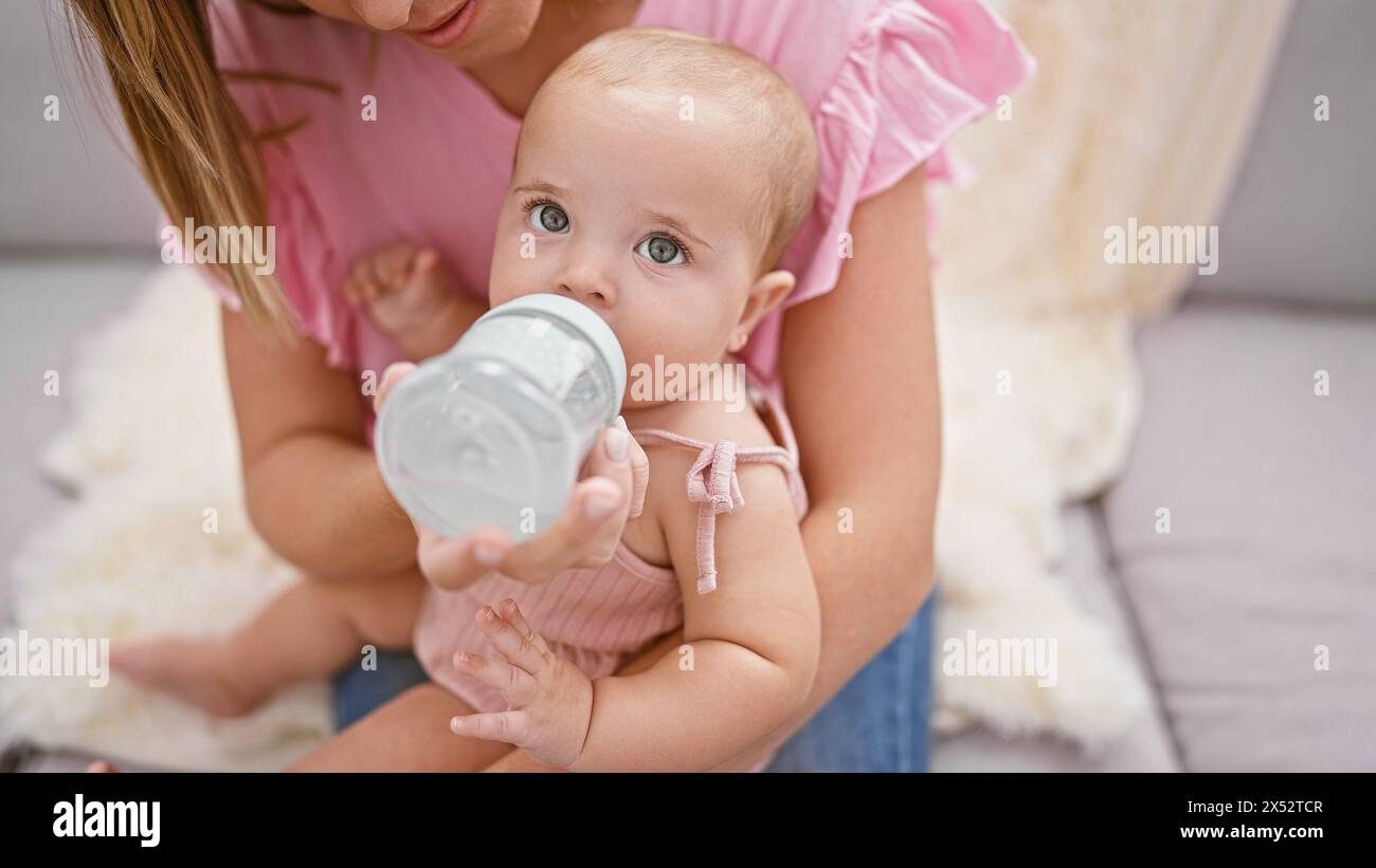 Loving mother giving a milk feeding bottle to her baby daughter while sitting in the relaxed comfort of their family home, cherishing an intimate indo Stock Photo