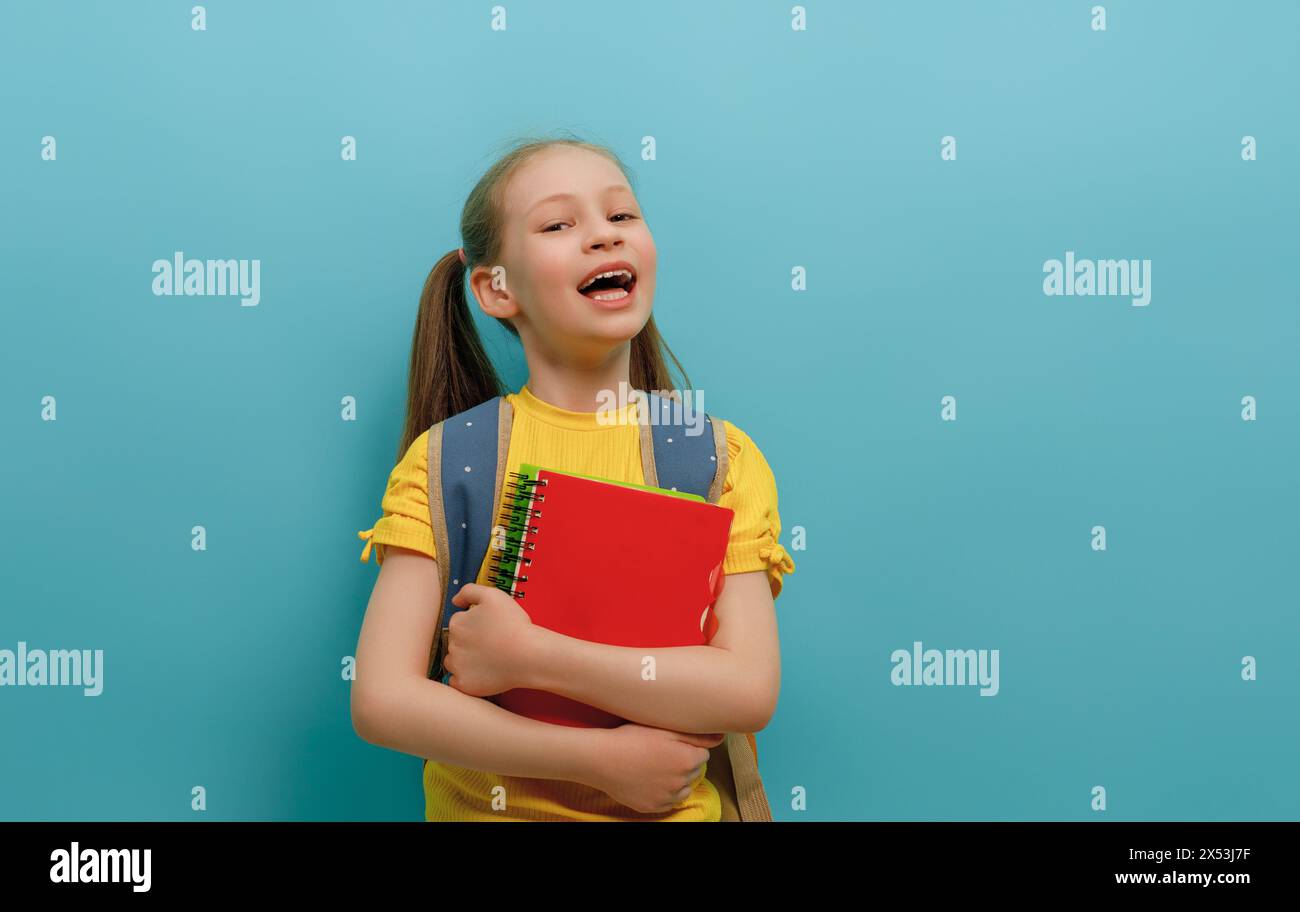 Cute happy kid standing on light blue background. Child with backpack, little girl is ready to back to school. Stock Photo