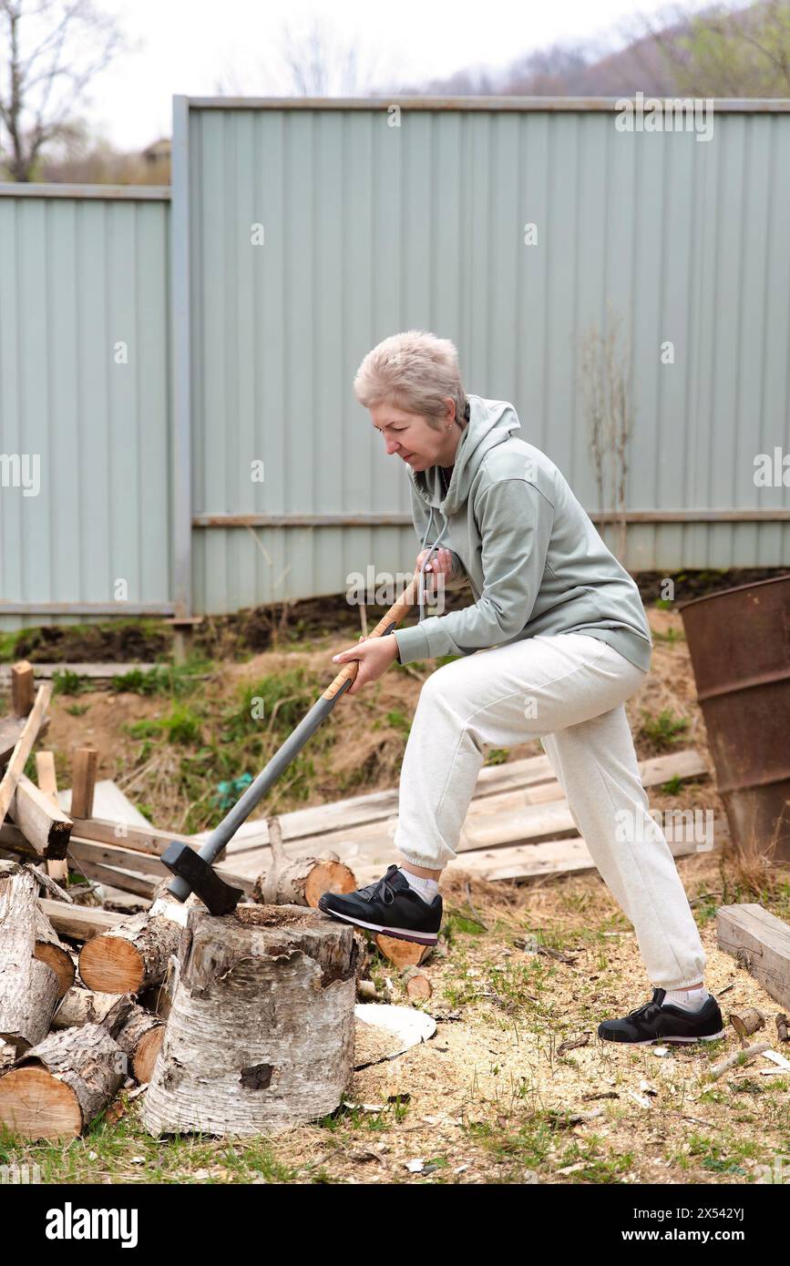 russian mature 57 y.o. woman in a tracksuit chopping wood Stock Photo