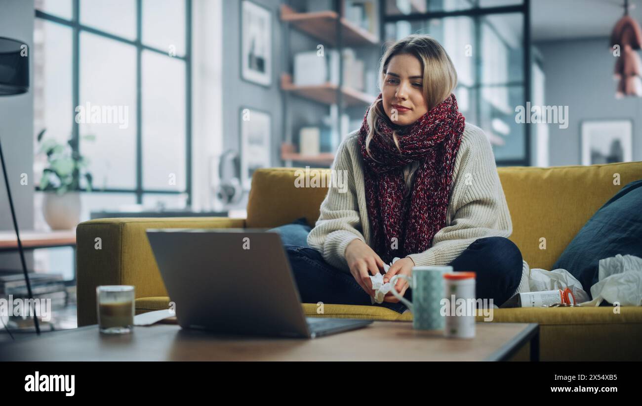 Ill Young Female Blowing Her Nose and Calling into Office with Video Phone Call from Cozy Living Room. Sick and Nauseous Woman in Warm Scarf is Using Laptop for Watching Movies. Stock Photo