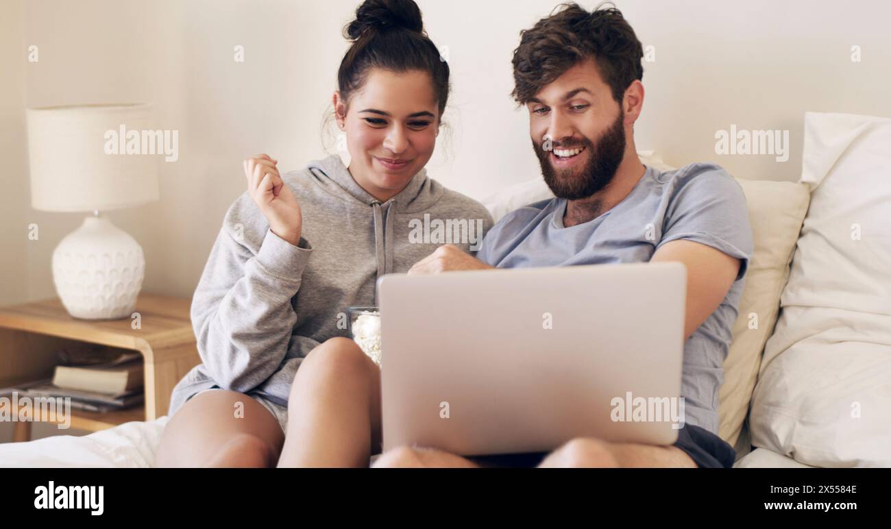 Happy, couple and relaxing with laptop on bed for bonding by watching movie, film and comedy together at house. Man, woman and care with popcorn in Stock Photo