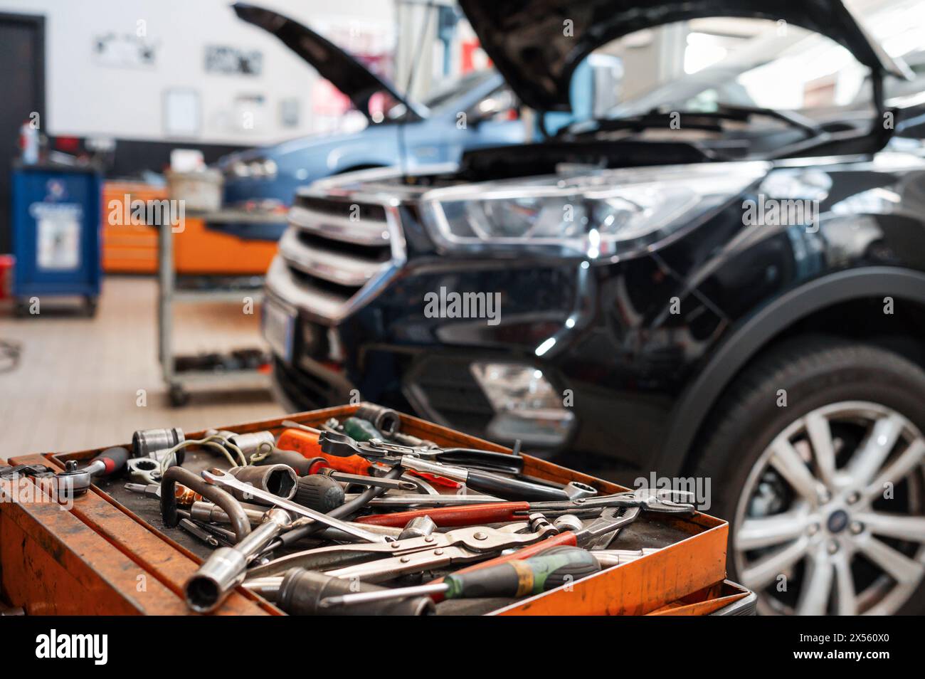 Various mechanical tools and instruments comprising screwdriver wrench plier placed in oily tray in front of car with opened bonnet and hanging headli Stock Photo