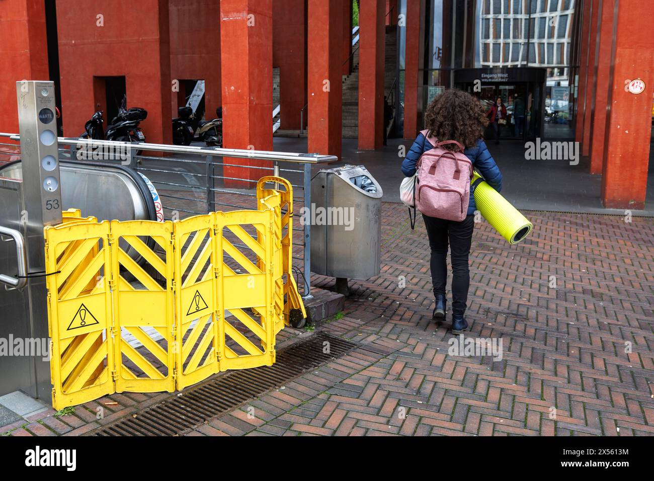a woman with a yellow yoga mat passes a yellow barrier at an escalator at the tonhall in the Deutz district of Cologne, Germany. eine Frau mit einer g Stock Photo
