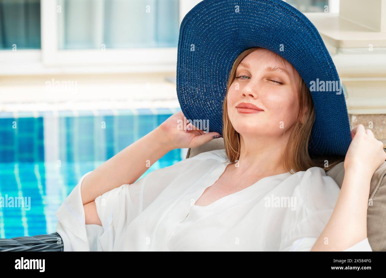 Young woman sitting near the pool winks her eyes in a blue hat Stock Photo