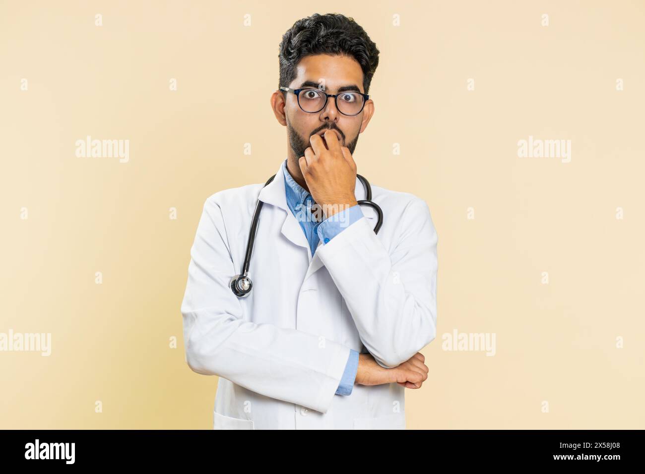 Upset scared frightened Indian doctor cardiologist man biting nails, feeling worried nervous about serious troubles, stress, anxiety disorder, panic attack, guilty. Arabian guy on beige background Stock Photo