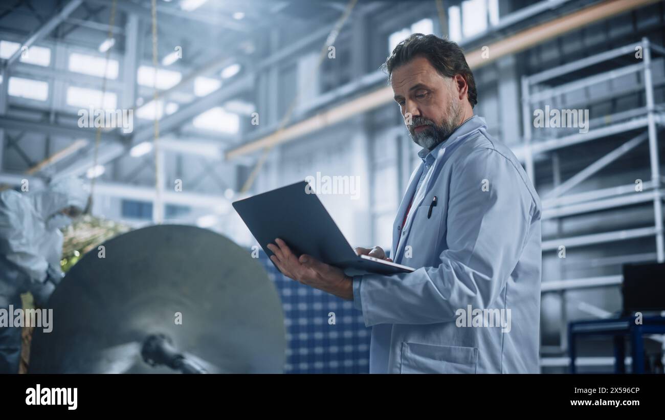 Male Engineer Uses Laptop Computer while Working on Satellite Construction. Aerospace Agency Manufacturing Facility: Scientists Develop, Assemble Spacecraft for Space Exploration, Observation Mission Stock Photo
