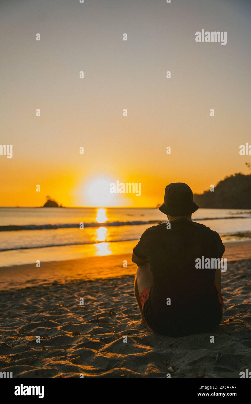 A person sits contemplatively on the beach, watching the sun set over ...