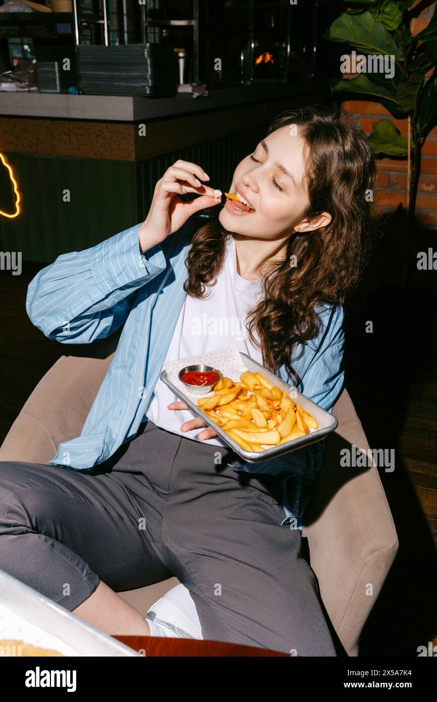 A woman enjoys a tray of golden French fries with a side of ketchup in a relaxed dining setting, embodying a casual and enjoyable mealtime. Stock Photo