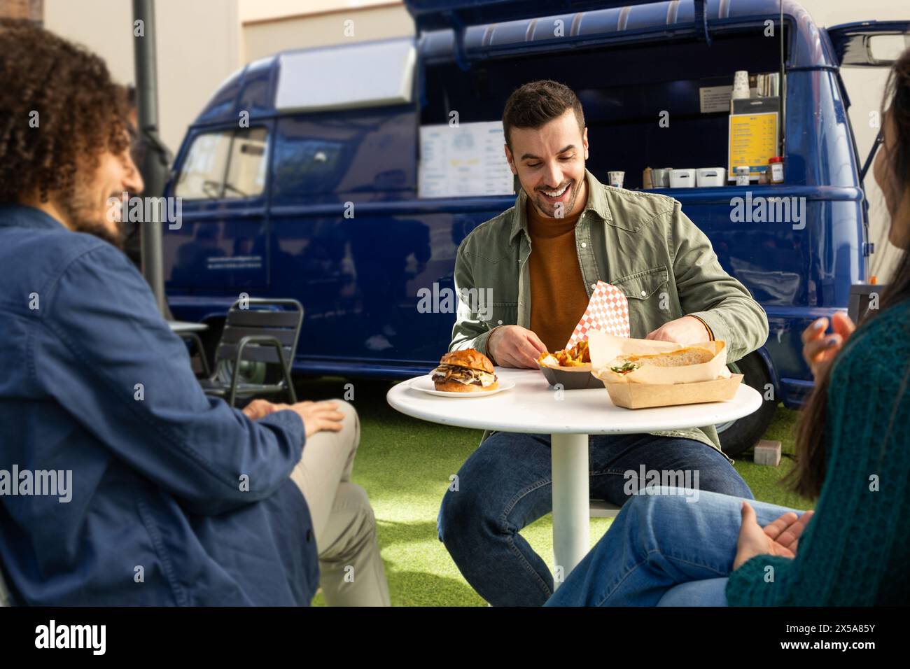 Friends share smiles and a casual meal outside a trendy blue food truck, showcasing an enjoyable urban eating experience Stock Photo
