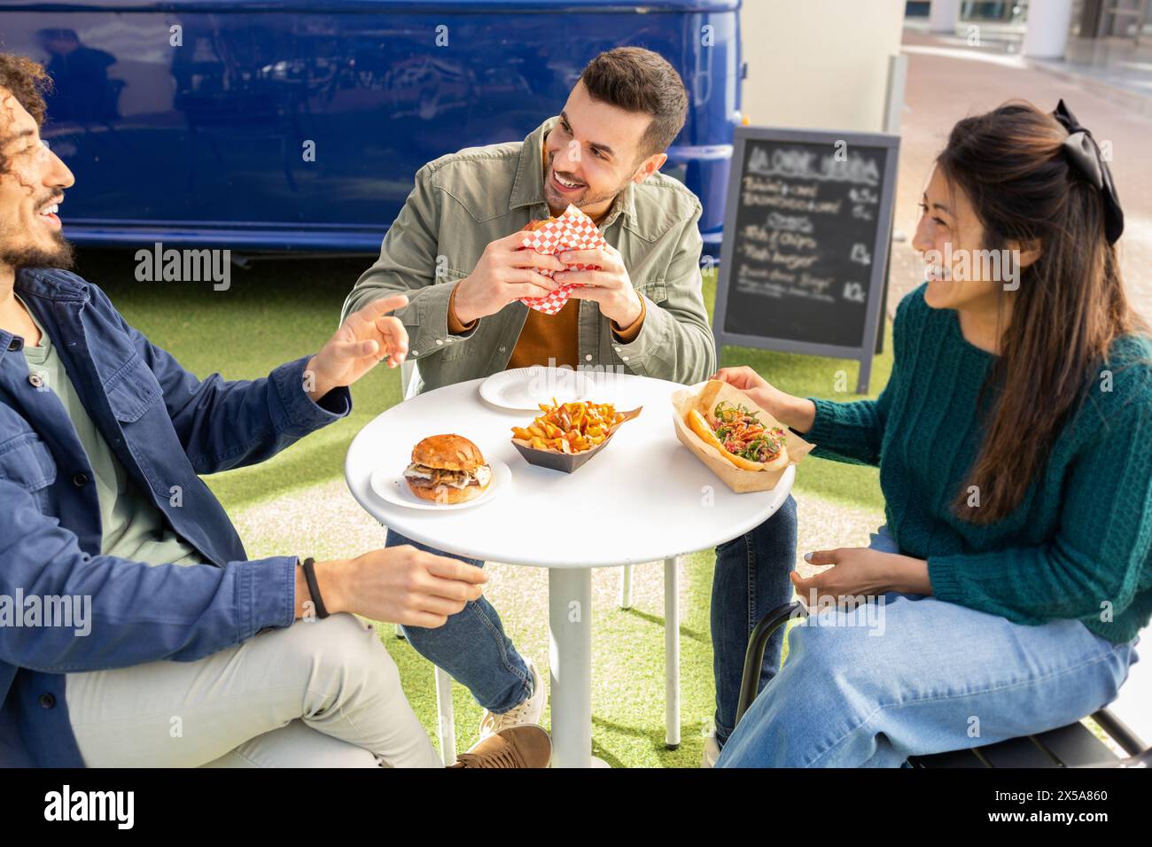 Three friends share a laugh while eating street food from a food truck, seated at an outdoor table in a casual setting Stock Photo