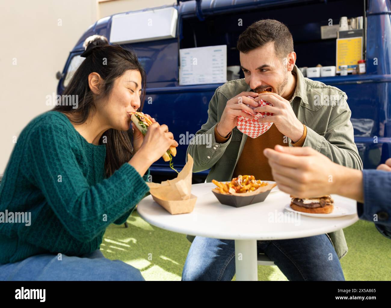 Two friends happily eating street food by a food truck, sharing a casual outdoor dining experience Stock Photo