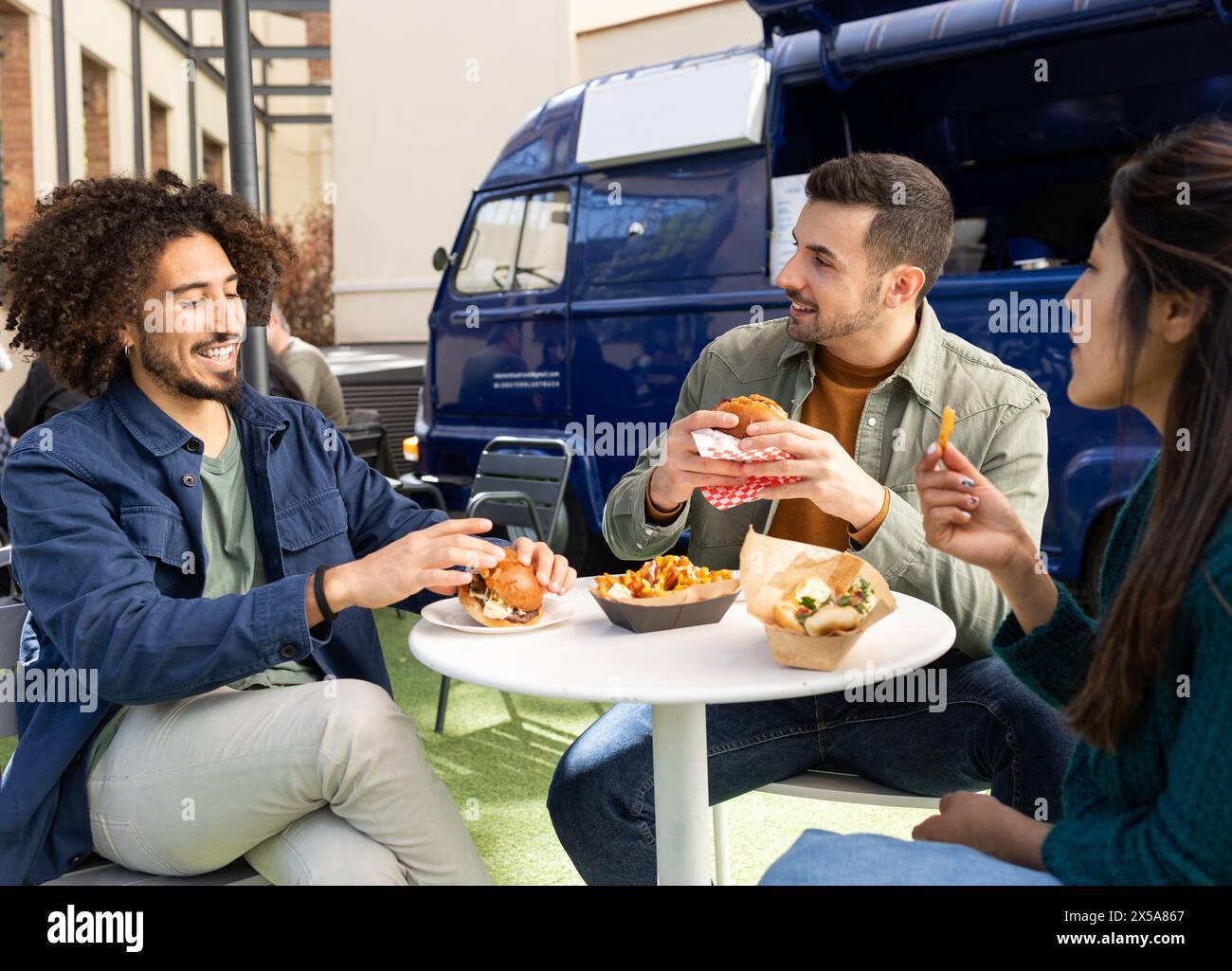 Three friends sit and laugh together while eating burgers and fries outside a blue food truck Stock Photo