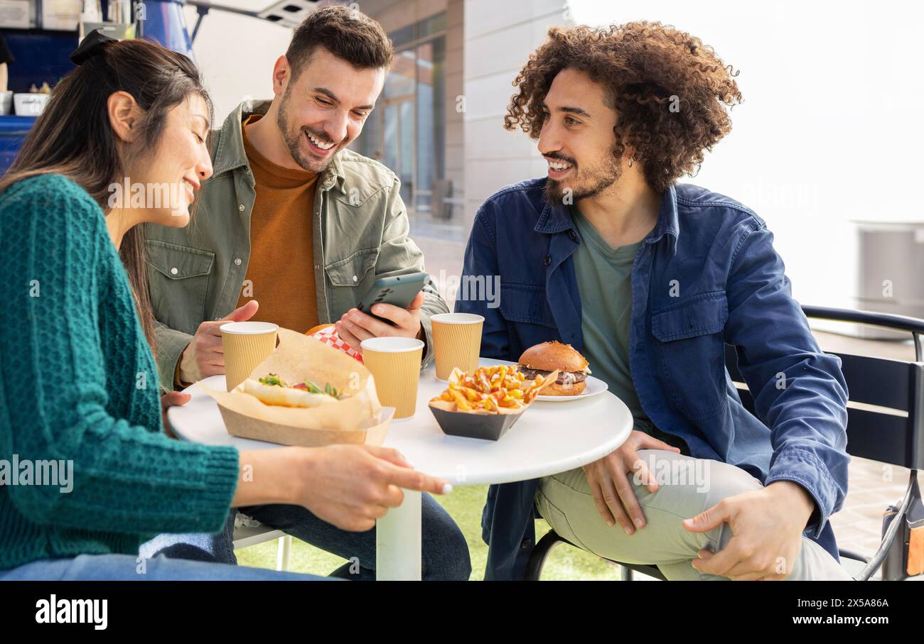 A group of happy friends enjoy a casual meal together, seated outside with food truck fare like burgers and fries Stock Photo