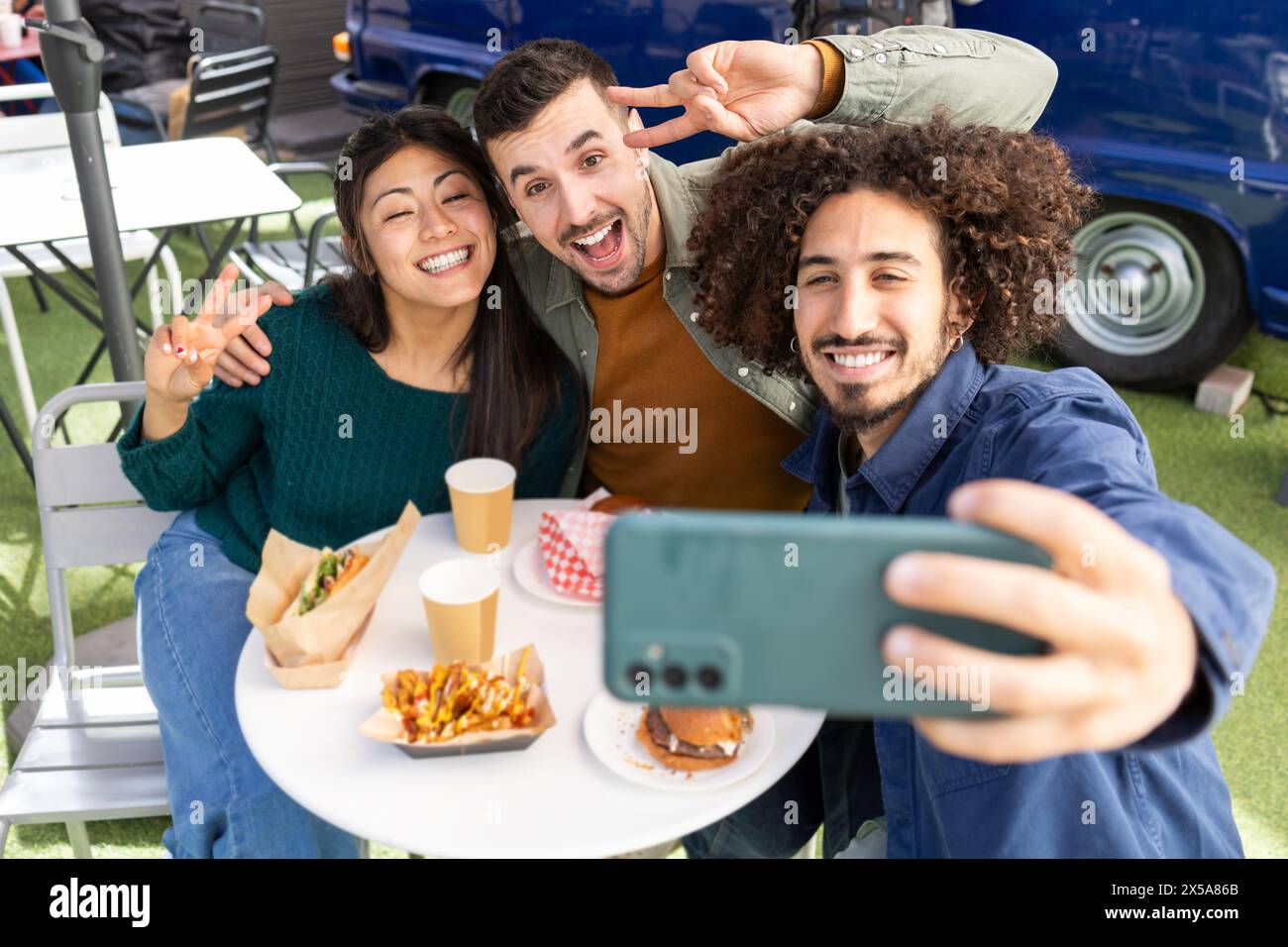 Three friends enjoy a meal from a food truck while taking a selfie, showcasing fun and friendship Stock Photo
