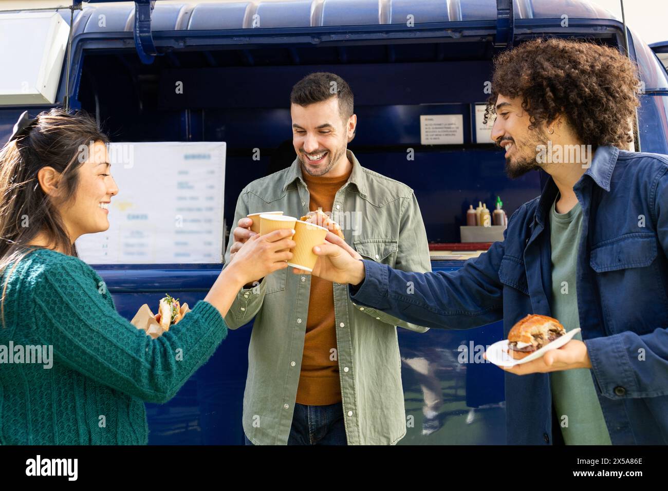 Three friends enjoy a sunny day, laughing and sharing street food near a vibrant blue food truck Stock Photo