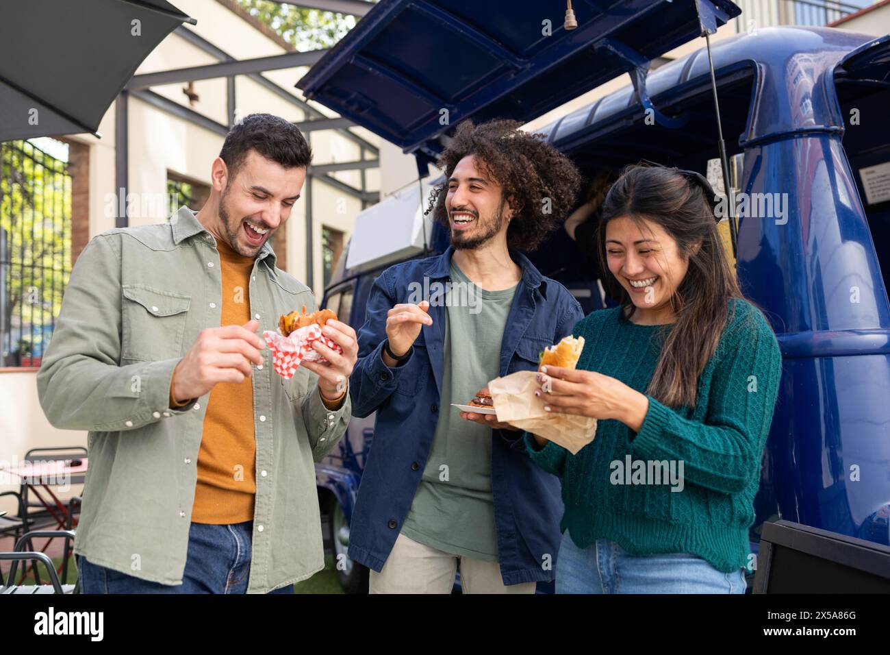 Three happy friends enjoy street food by a vibrant blue food truck, laughing and sharing a sunny moment together Stock Photo