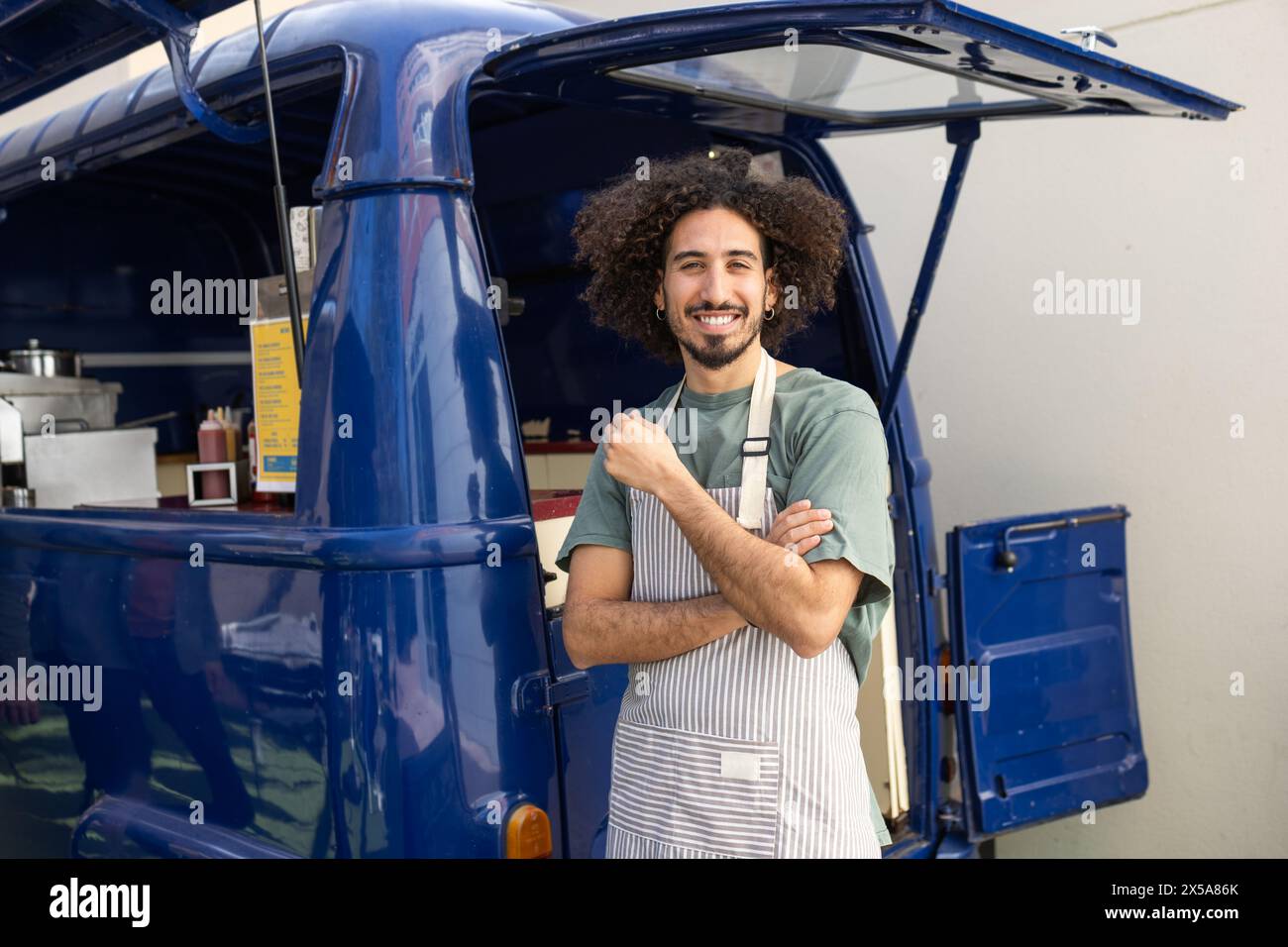 A jovial man with curly hair stands confidently outside his blue food truck, sporting a striped apron and giving a thumbs up Stock Photo