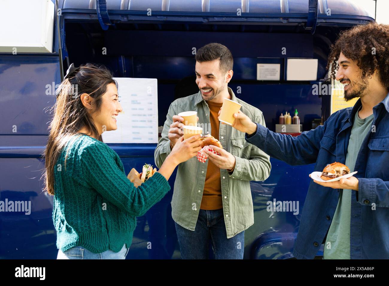 Three friends share a moment of joy while eating street food and drinking coffee beside a blue food truck under a bright, clear sky Stock Photo