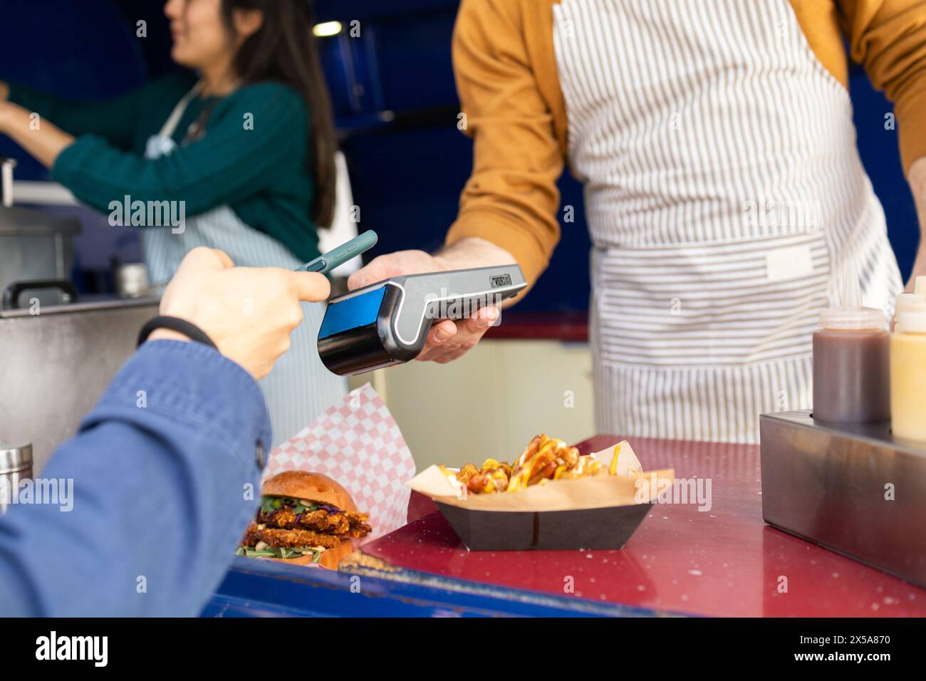 Customer making a card payment for street food at a busy food truck while friends work together in the background Stock Photo