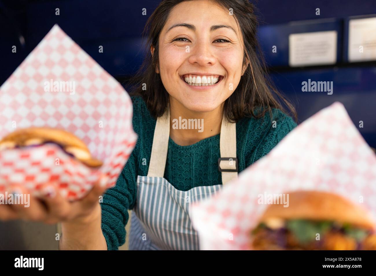 Smiling woman in a food truck holding out delicious burgers wrapped in paper, exuding enthusiasm and friendliness Stock Photo