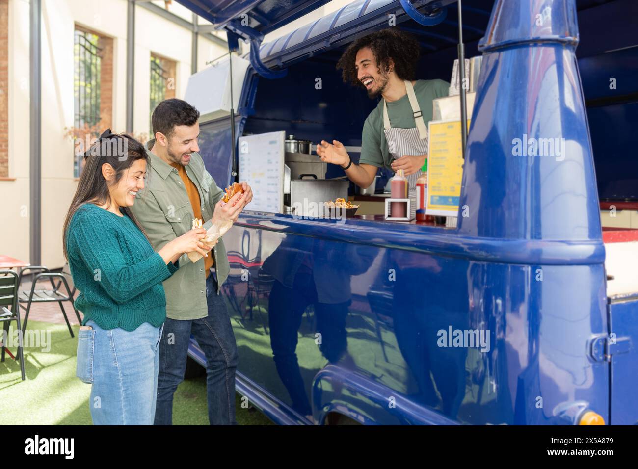 A group of friends share a laugh while purchasing fast food from a cheerful vendor inside a vibrant blue food truck Stock Photo