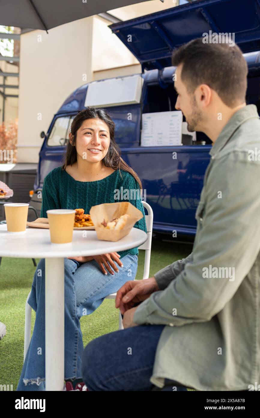 Two friends enjoying a casual lunch seated at a table outside a blue food truck, with one person eating and both holding cups Stock Photo