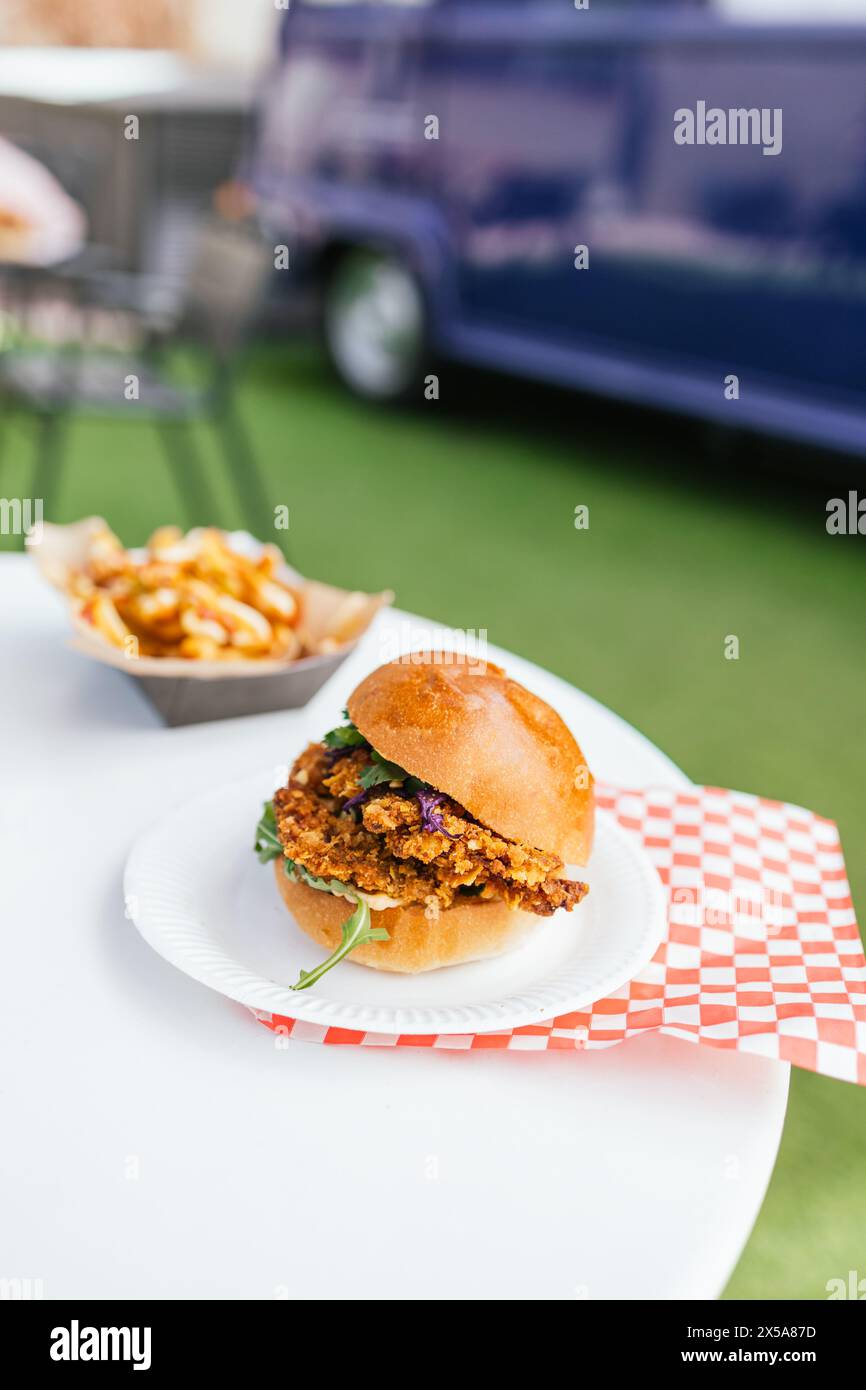 A close-up image of a delicious gourmet burger paired with seasoned fries served on a checkered paper, with a food truck in the background Stock Photo