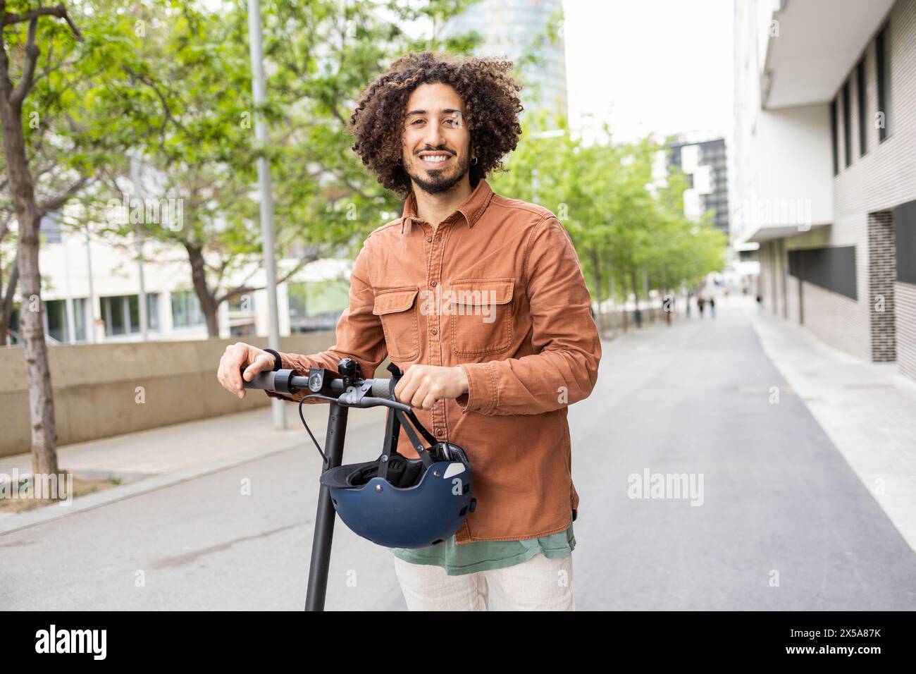 Confident young man with curly hair, smiling while riding an electric scooter in a modern urban environment, holding a helmet Stock Photo