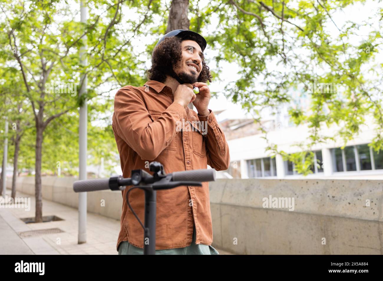 A cheerful man adjusts his helmet in preparation to ride his electric scooter, standing in an urban park lined with lush trees Stock Photo