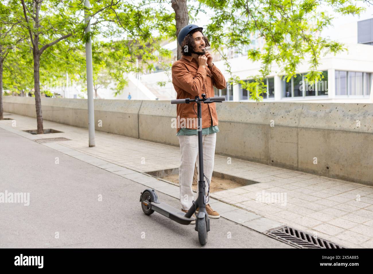 A young man in casual attire stands with an electric scooter, holding his chin thoughtfully in an urban, tree-lined area Stock Photo