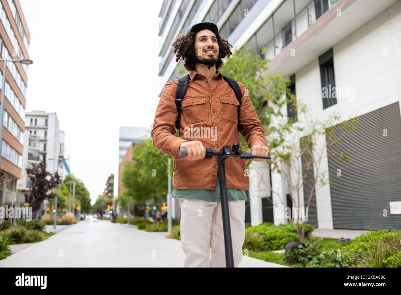 Cheerful curly-haired man navigates through a modern urban environment on an electric scooter Dressed in a stylish brown jacket and carrying a backpac Stock Photo