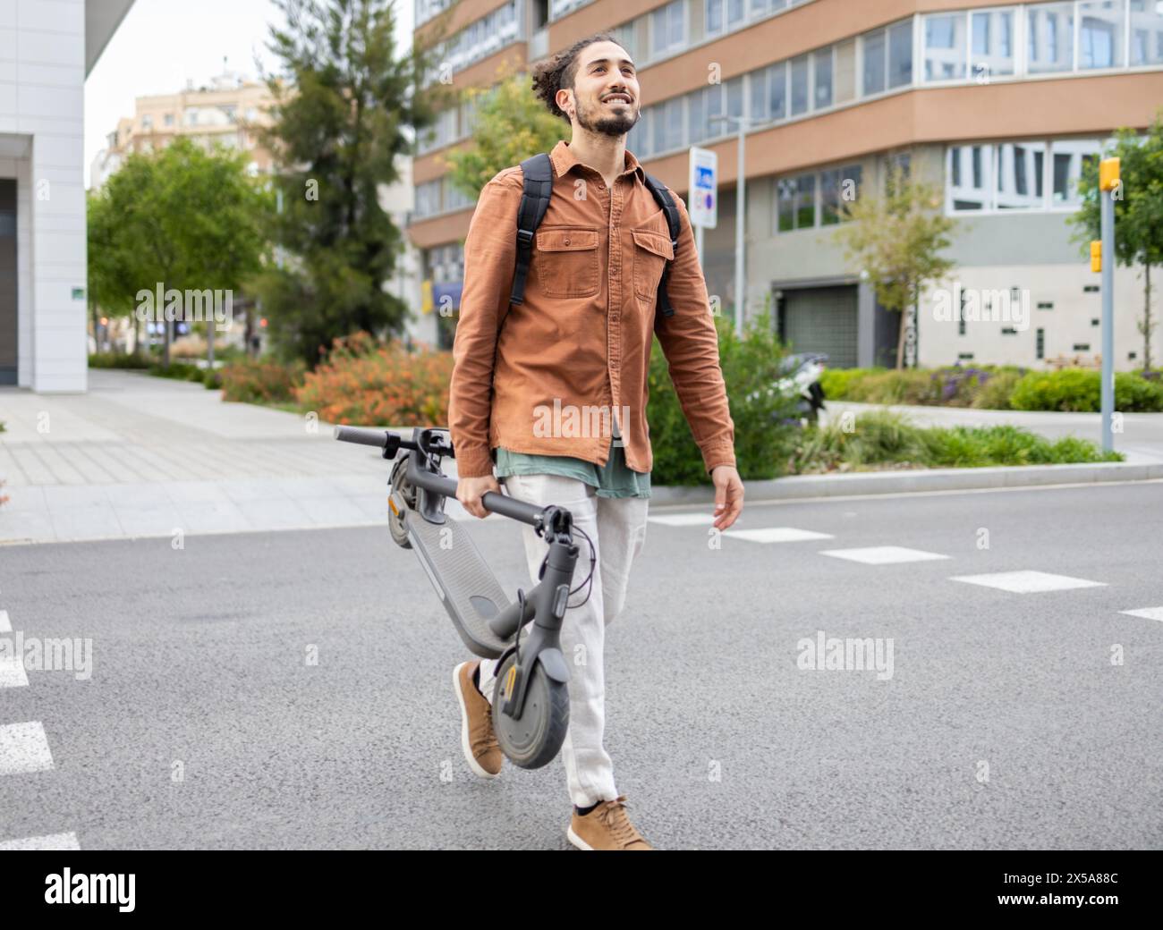 A young man is captured walking cheerfully with his electric scooter in hand, on a modern city sidewalk, epitomizing urban mobility and a sustainable Stock Photo