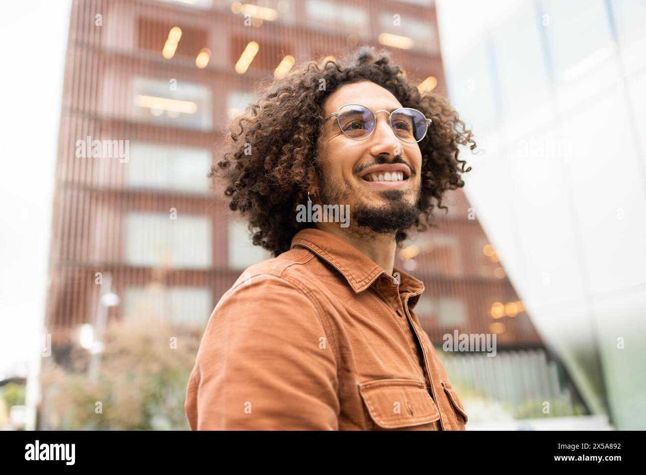 Joyful young man with curly hair rides an electric scooter through an urban area, expressing freedom and happiness against a modern building backdrop Stock Photo