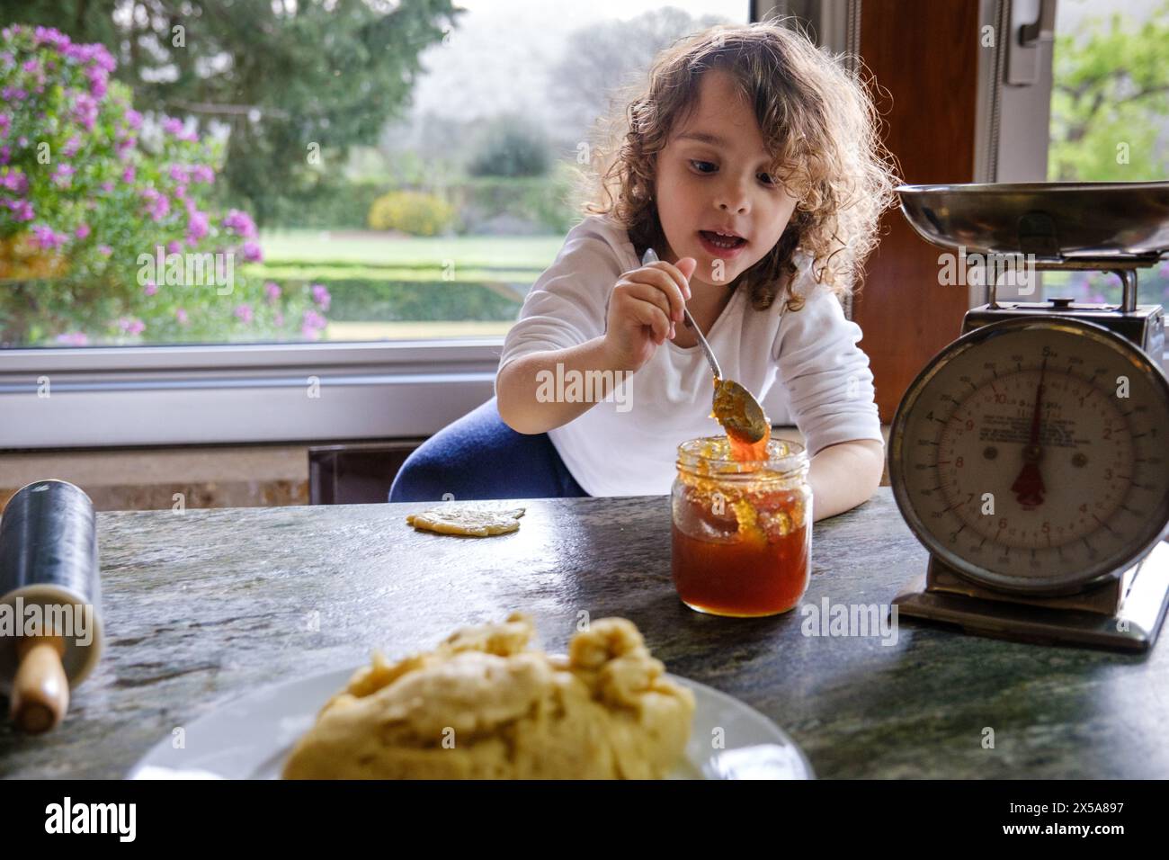 Curious child baking in a home kitchen with dough, a rolling pin, and jam next to a vintage scale, with a garden view window Stock Photo