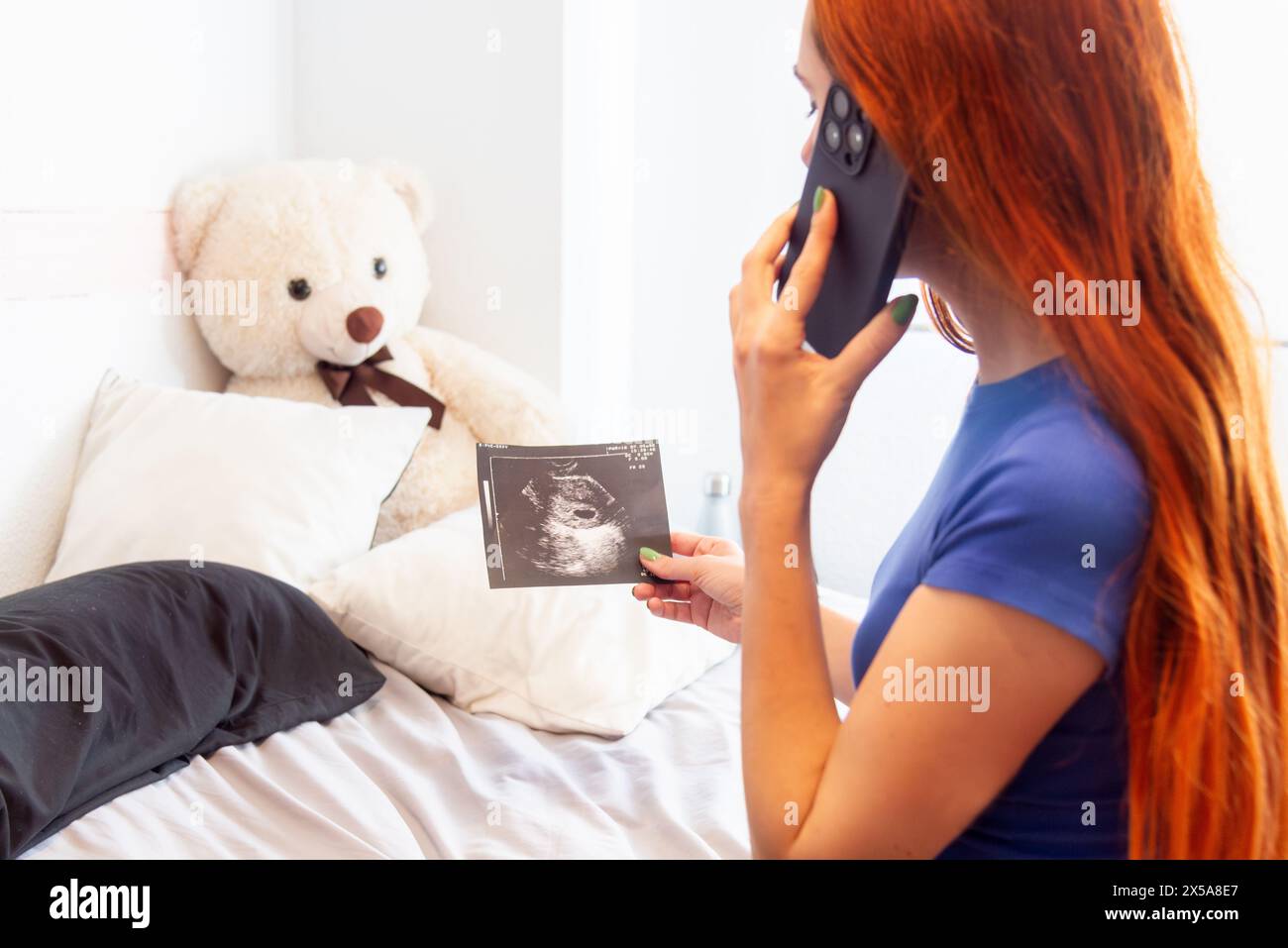 A young pregnant woman with red hair is chatting on her mobile phone, excitedly sharing the news of her pregnancy as she holds an ultrasound image in Stock Photo