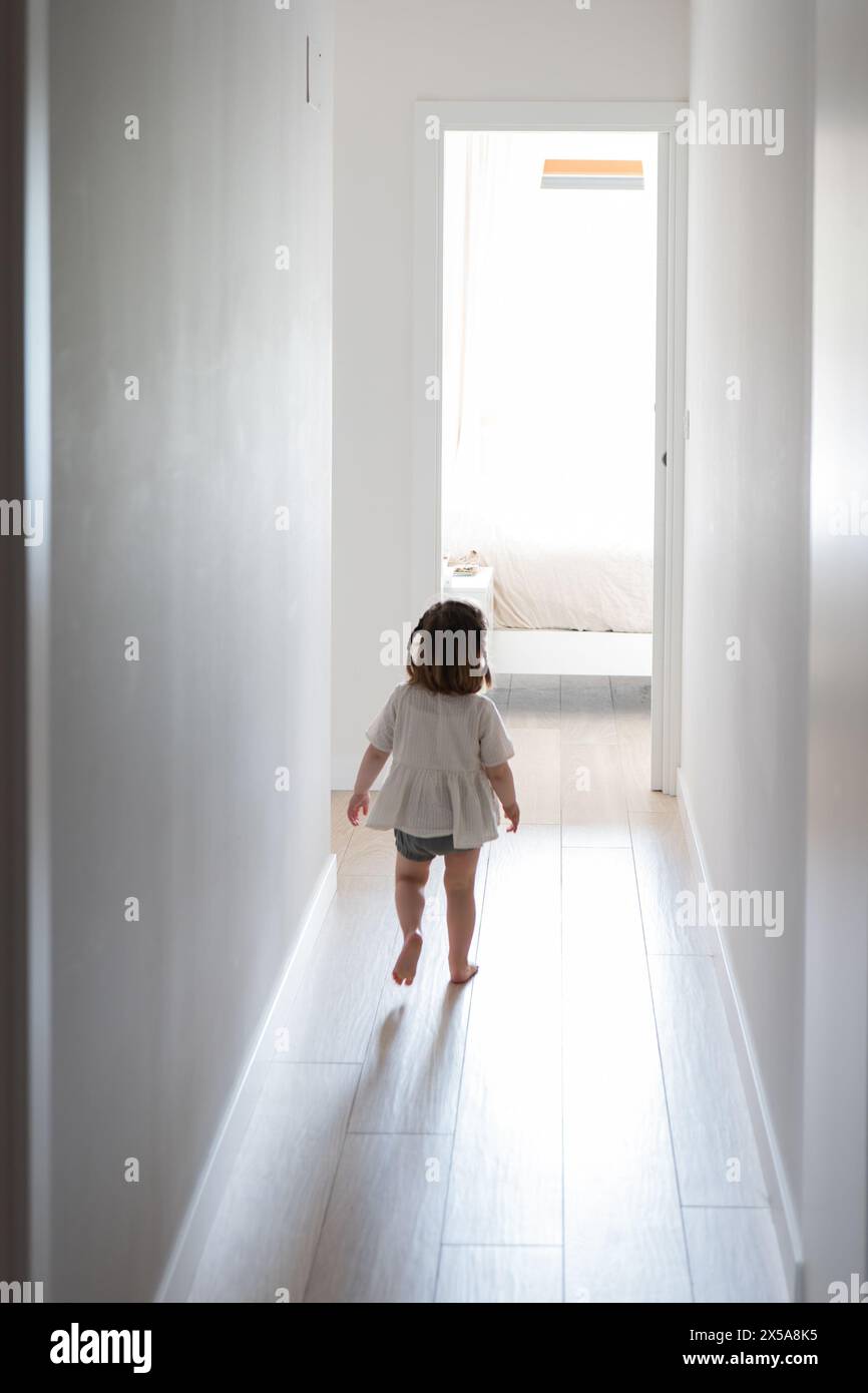 A toddler girl wanders barefoot down a bright hallway toward an open door with soft natural light spilling in Stock Photo