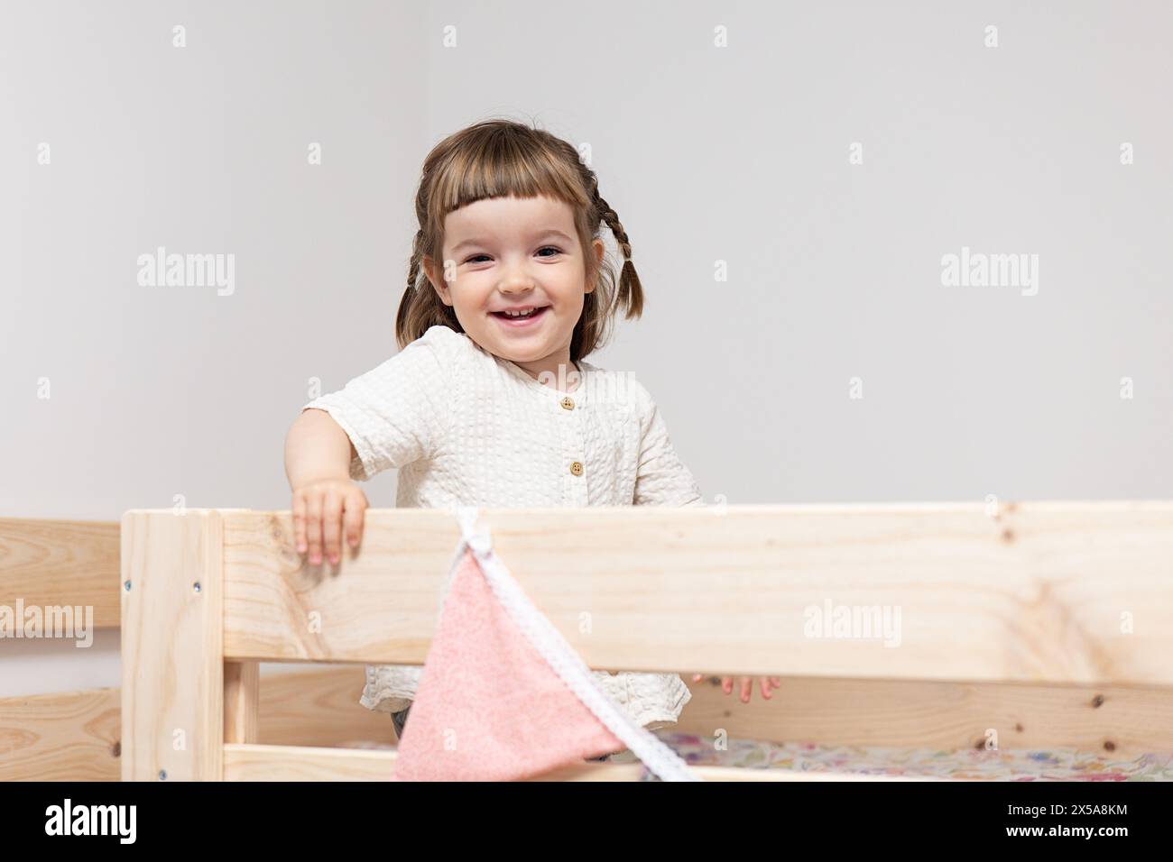 A cheerful toddler girl with pigtails smiles brightly while standing in a wooden bed with soft blankets Stock Photo