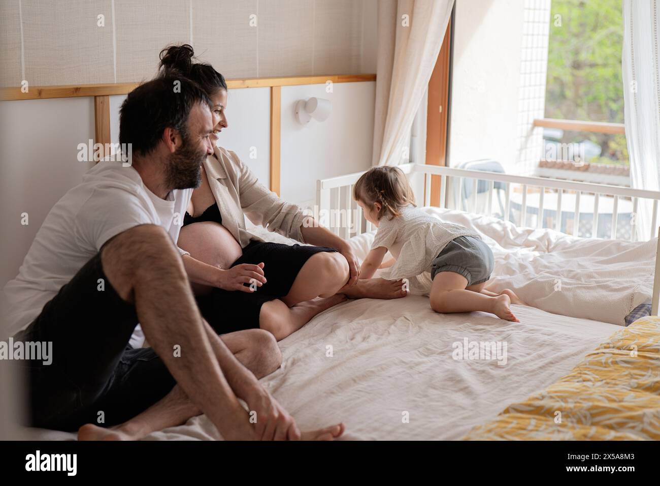 A heartwarming scene of a family with a toddler climbing on a bed near her pregnant mother and father, enjoying a relaxed moment together Stock Photo