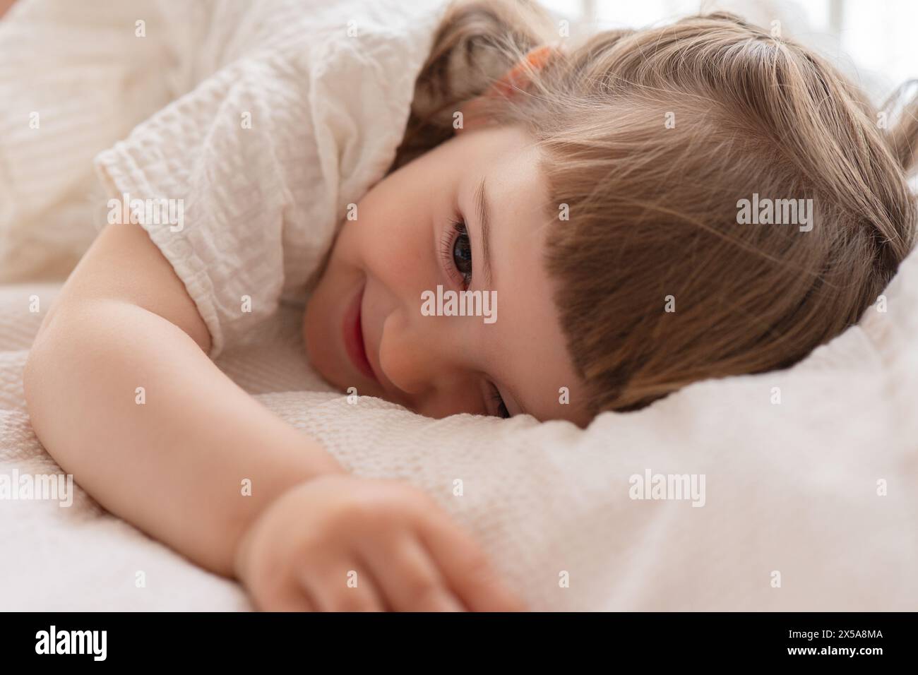 A young child with bright eyes and a gentle smile lies comfortably on a creamy textured blanket, radiating joy and innocence Stock Photo