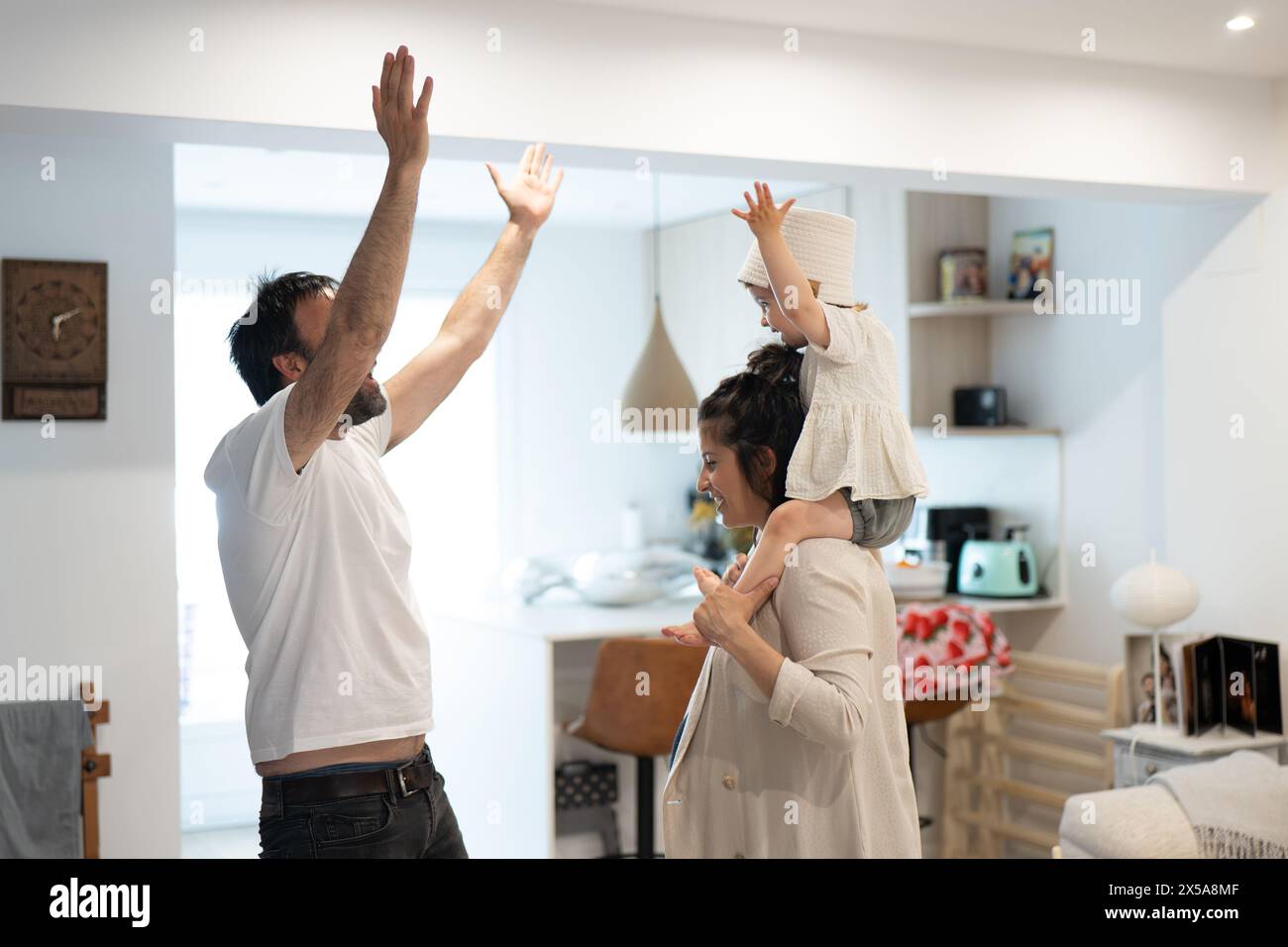 A young family enjoys a moment of play at home, with a child riding on his pregnant mothers shoulders and the father extending his hand in a gesture o Stock Photo