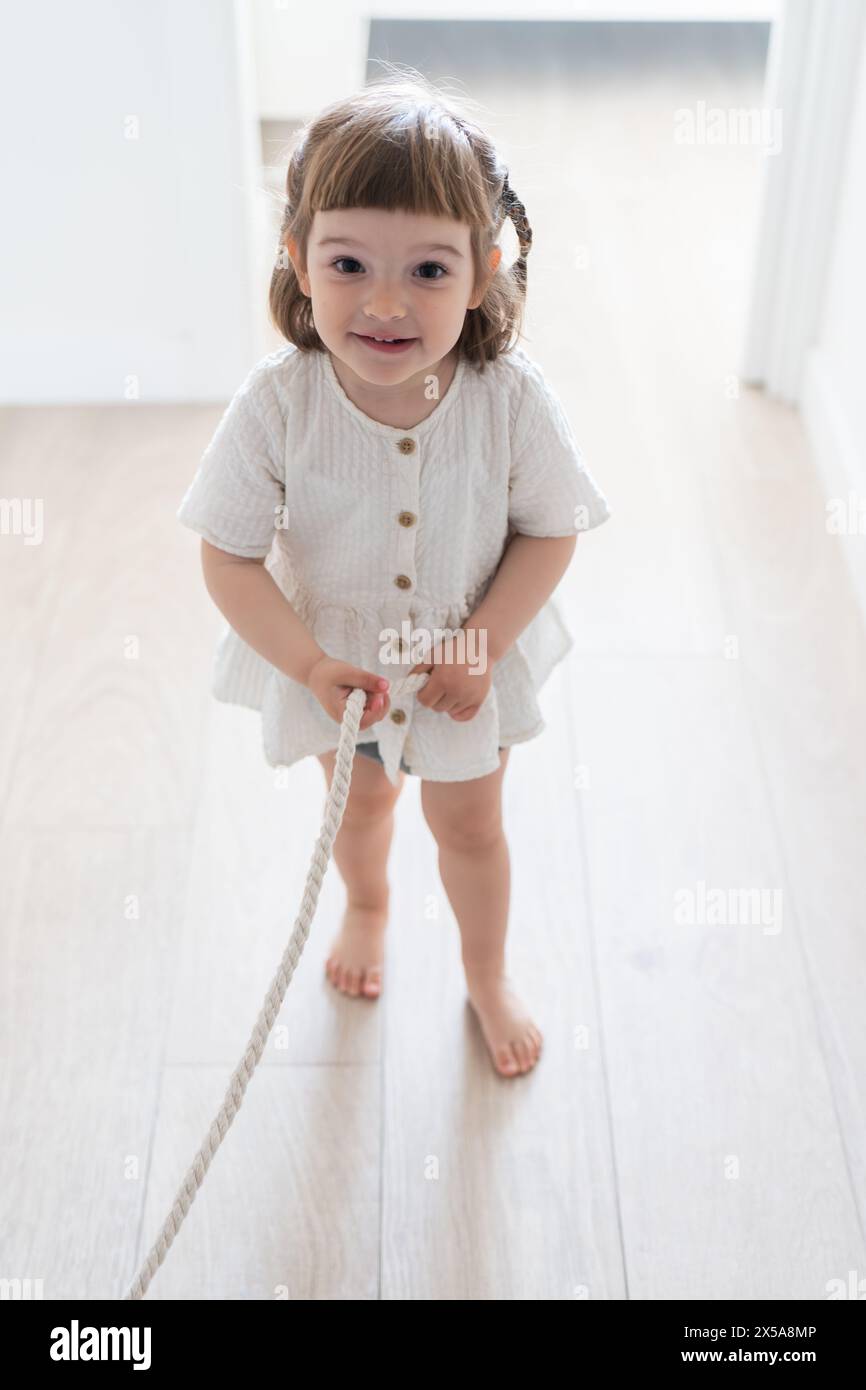 A cheerful toddler girl in a beige dress smiles while holding a rope inside a bright, minimalist room Stock Photo