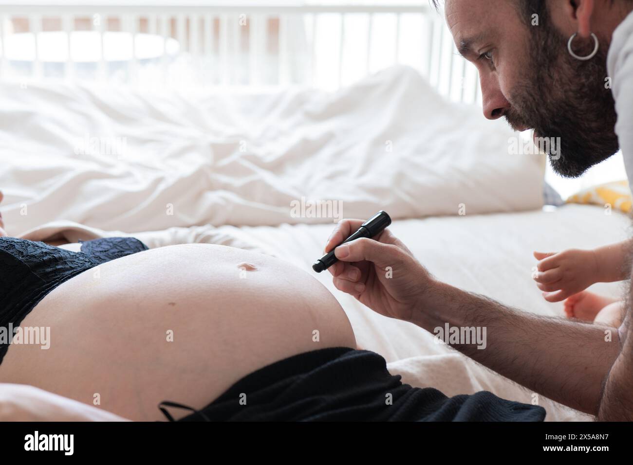 A soon-to-be father engages in a tender moment, drawing on his pregnant partners exposed belly while they sit on a bed Stock Photo