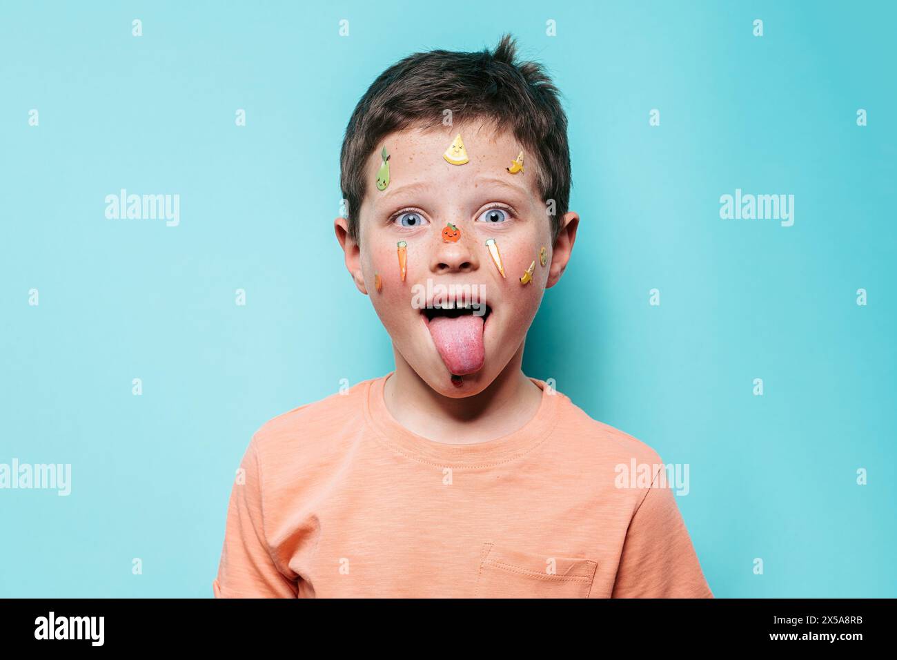 A young boy sticks out his tongue, face adorned with vibrant fruit stickers, against a teal background Stock Photo