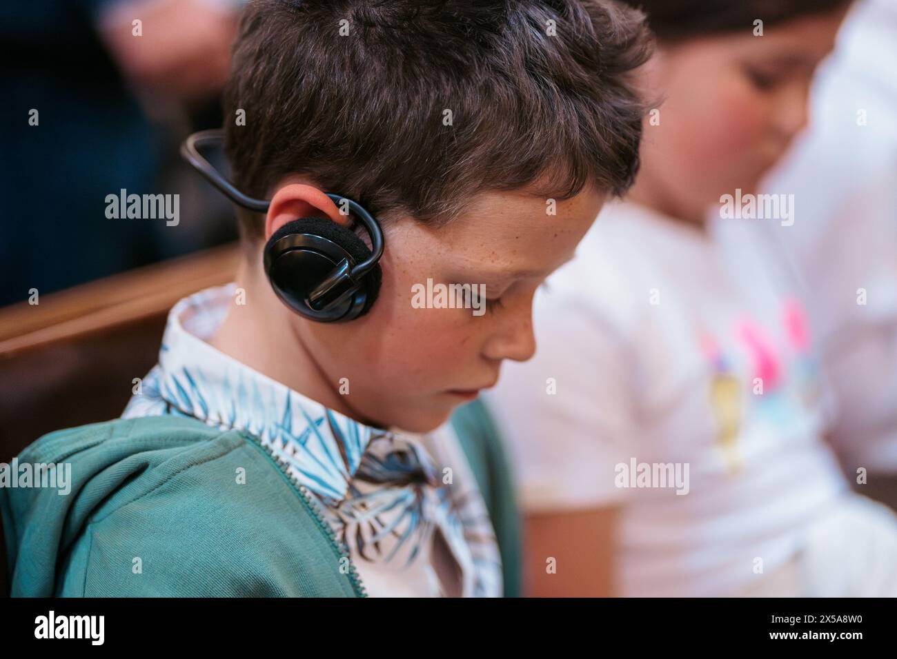 A young boy wearing a stylish bow tie and headphones is focused in a summer classroom atmosphere, with other children around him. Stock Photo