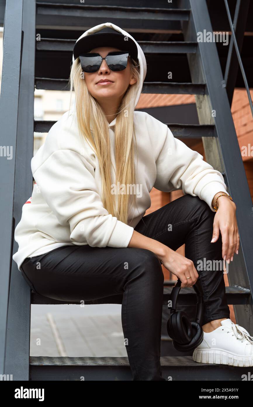 A stylish blonde woman in casual wear sits on metal stairs with sunglasses and headphones. Stock Photo