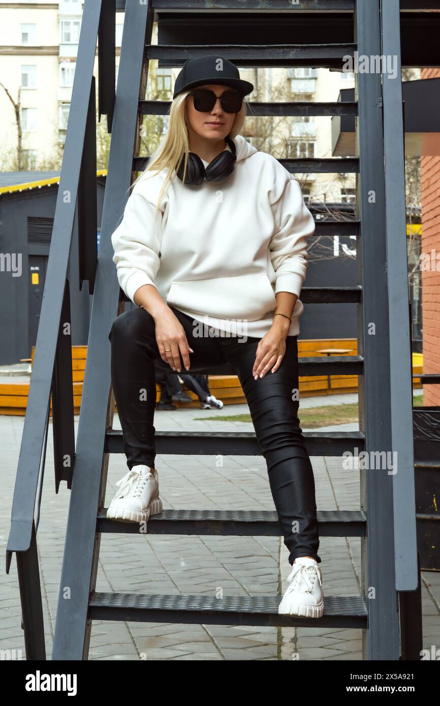 A fashionable blonde lady relaxes on a metal stairway in the city, sporting chic sunglasses and a trendy black hat. Stock Photo