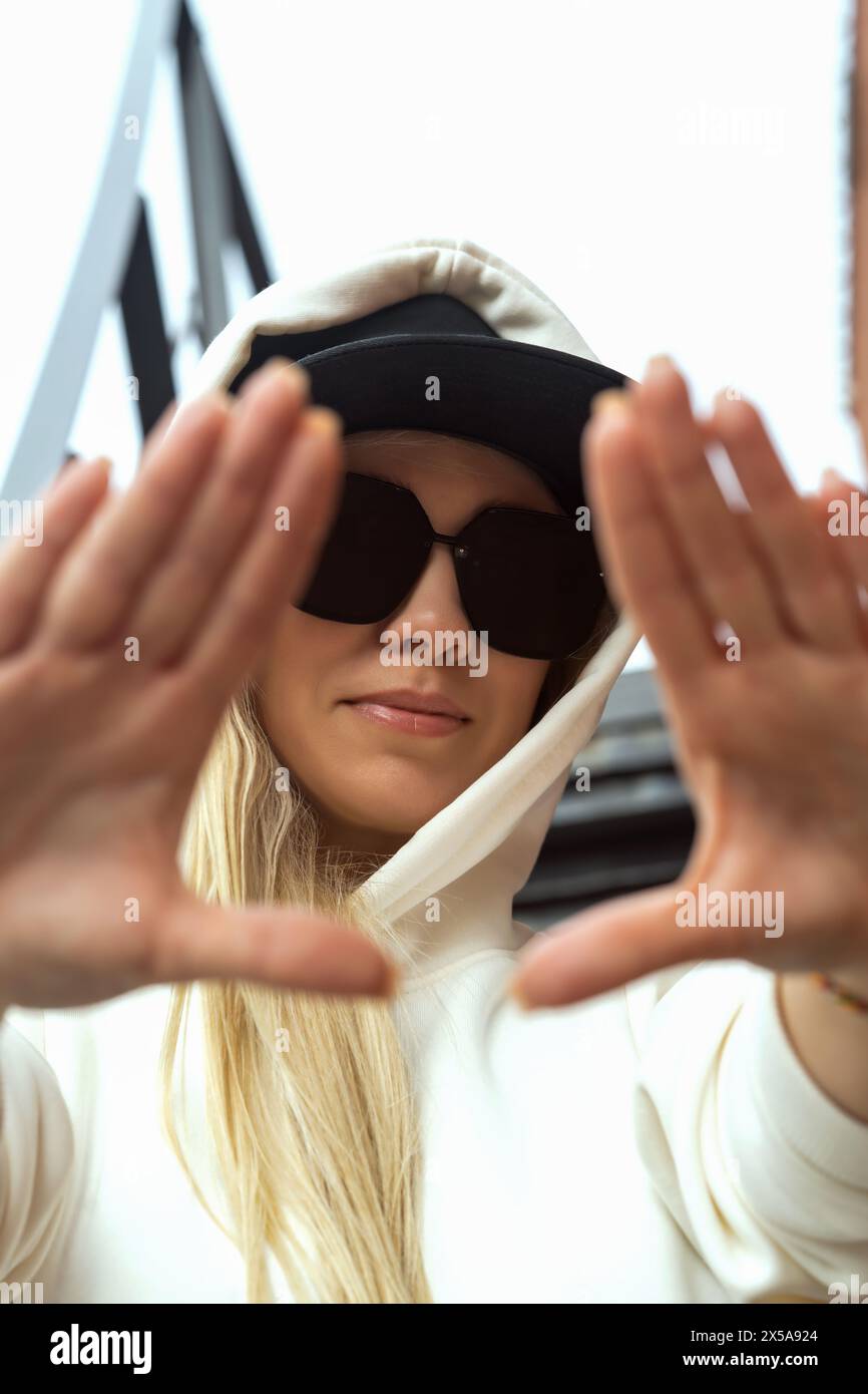 A stylish blonde woman in a hoodie and sunglasses frames her face with her hands on an urban street. Stock Photo