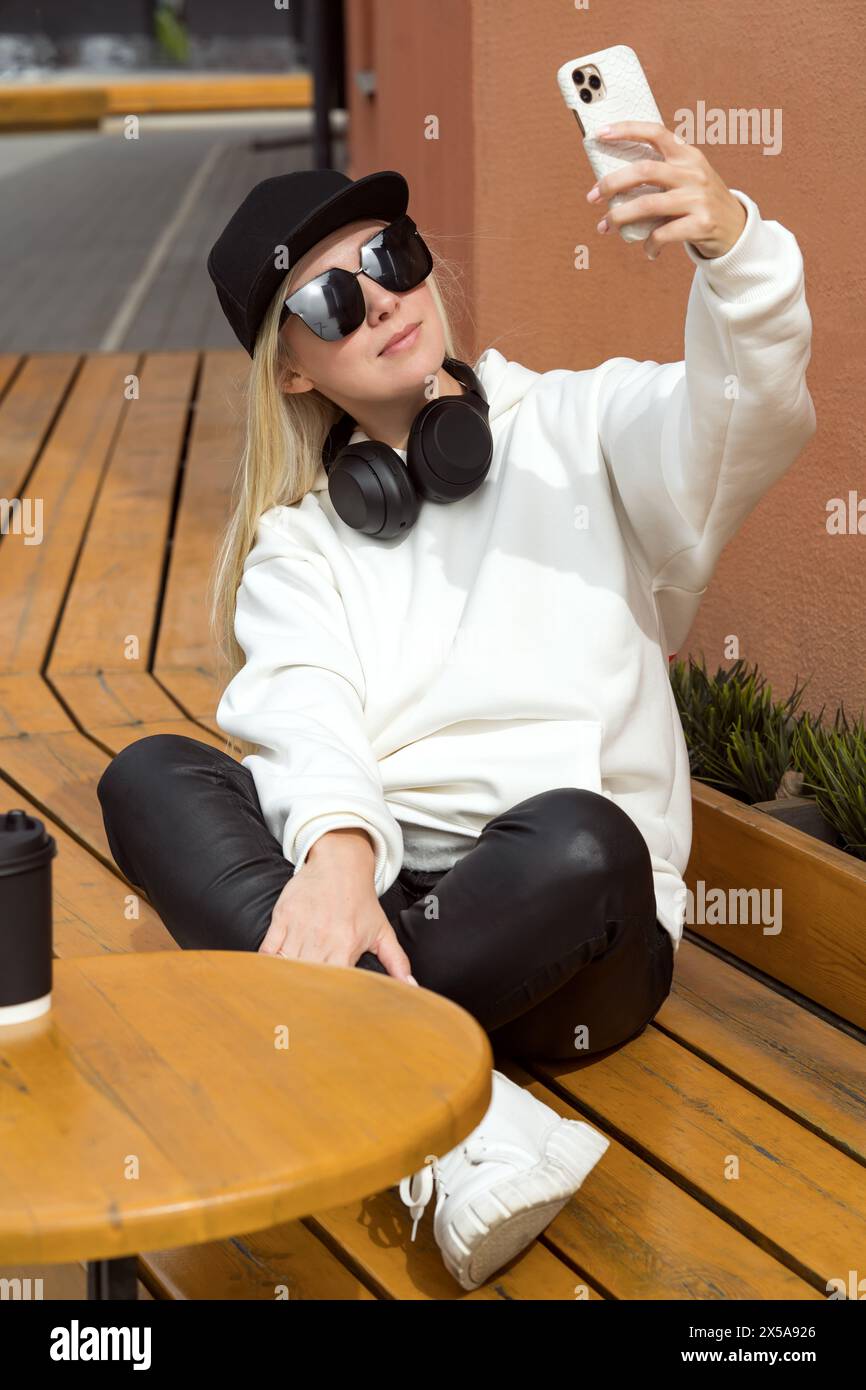 A stylish blonde woman in casual attire takes a selfie with her smartphone while sitting at a café terrace. Stock Photo