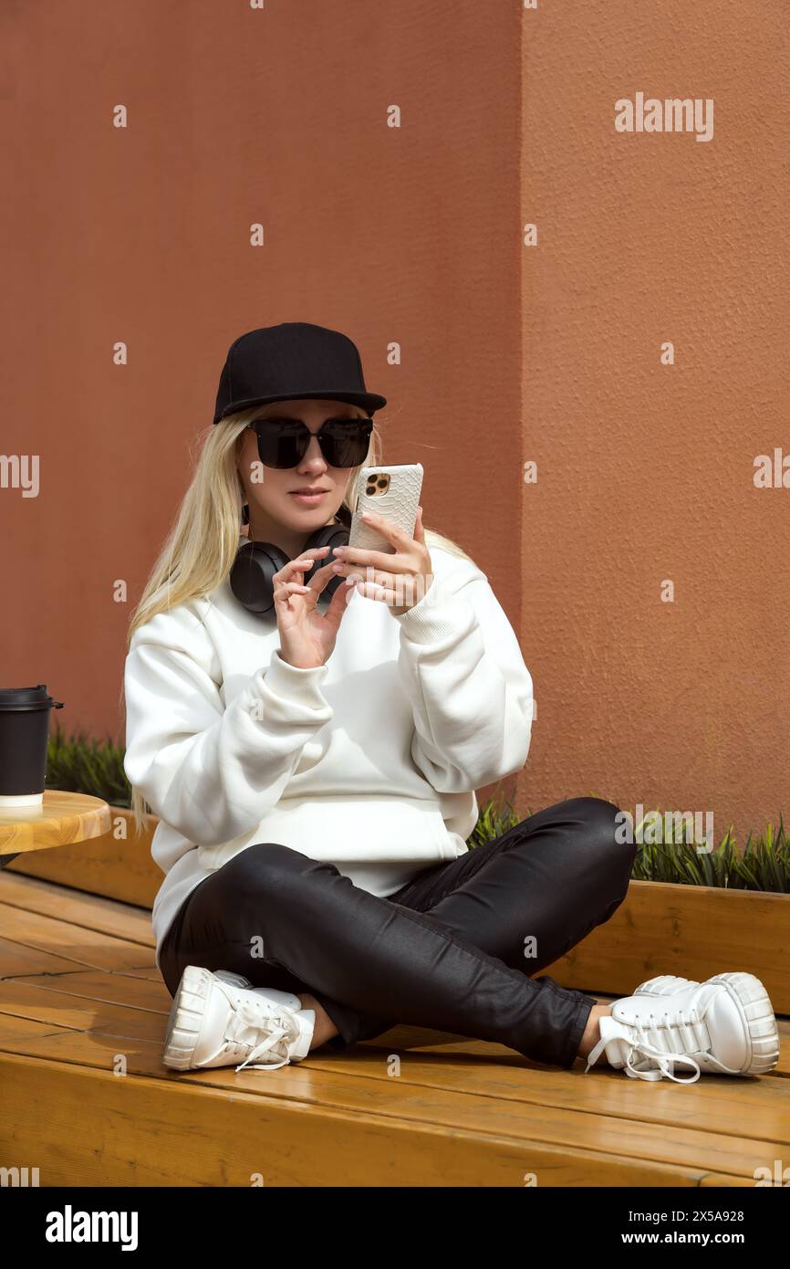 A trendy blonde woman in sunglasses and a black hat is engaged with her smartphone while sitting on wooden steps outdoors, with a coffee beside her. Stock Photo