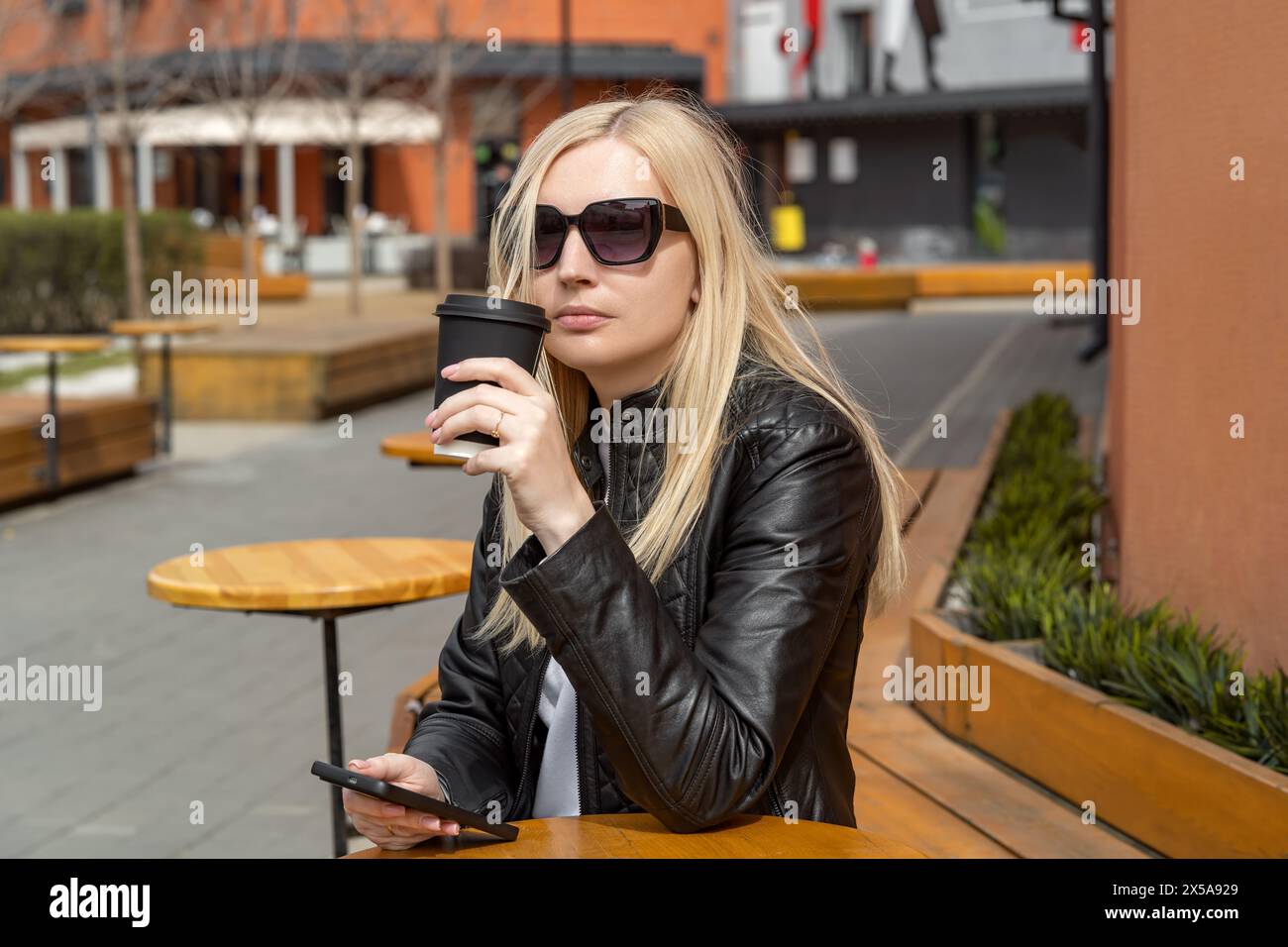 A fashionable woman in a leather jacket sips coffee while using her smartphone at an outdoor café Stock Photo